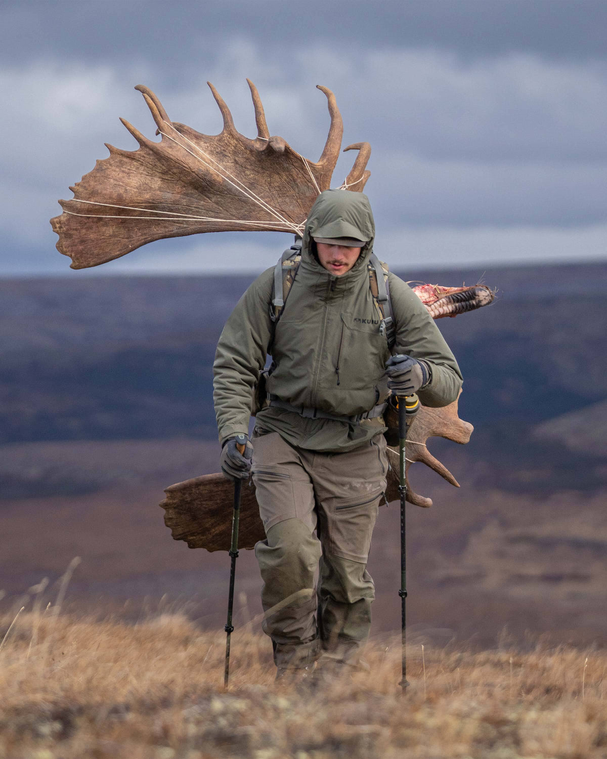 Hunter trekking up a hill carrying a large, palmated moose antlers on his back, wearing Mountain PRO Hooded Jacket in Ash Brown