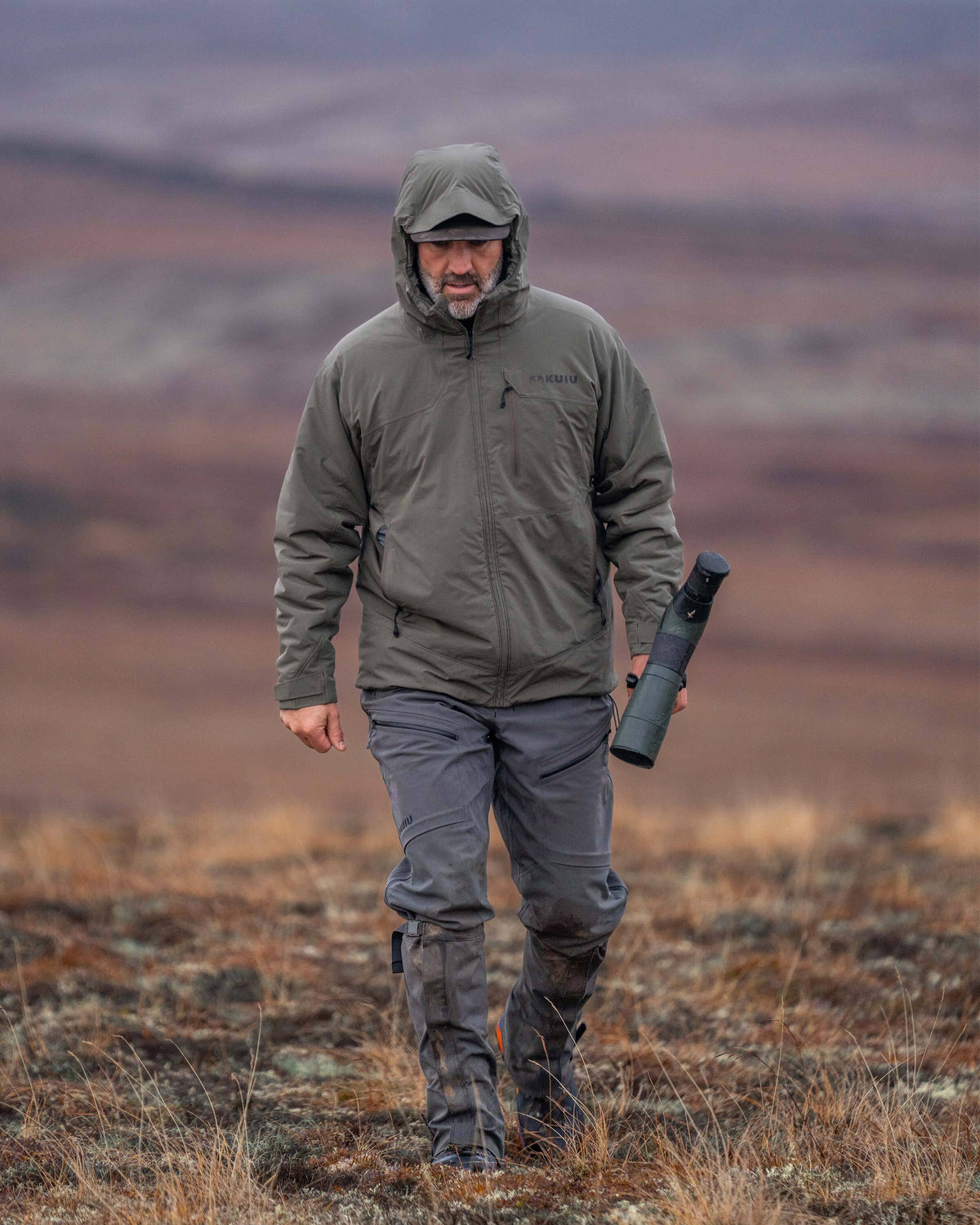 Hunter walking through a dry, low-shrub landscape while carrying a spotting scope wearing Moutain PRO Hooded Jacket in Ash Brown
