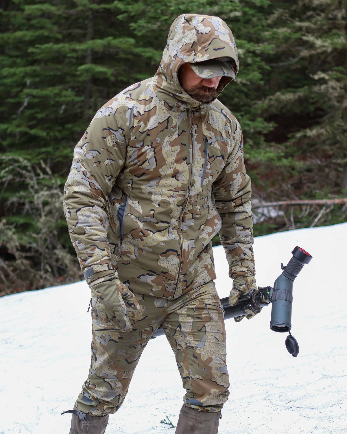 Hunter wearing Mountain PRO Hooded Jacket in Valo Camouflage, walks through the snow carrying a spotting scope and tripod