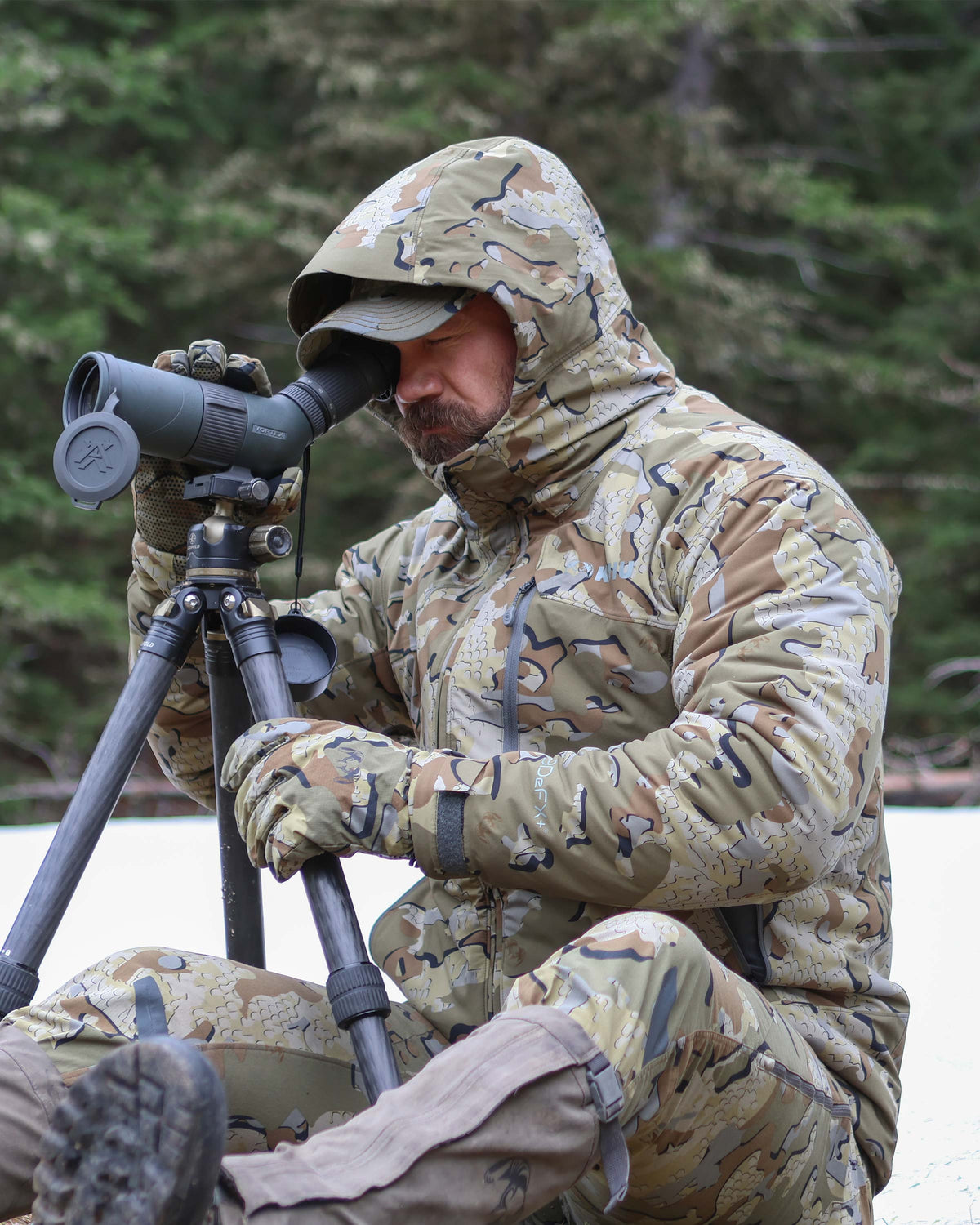 Hunter in a snowy forest setting, wearing Mountain PRO Hooded Jacket in Valo Camouflage, sitting on snow-covered ground, looking through a tripod-mounted spotting scope