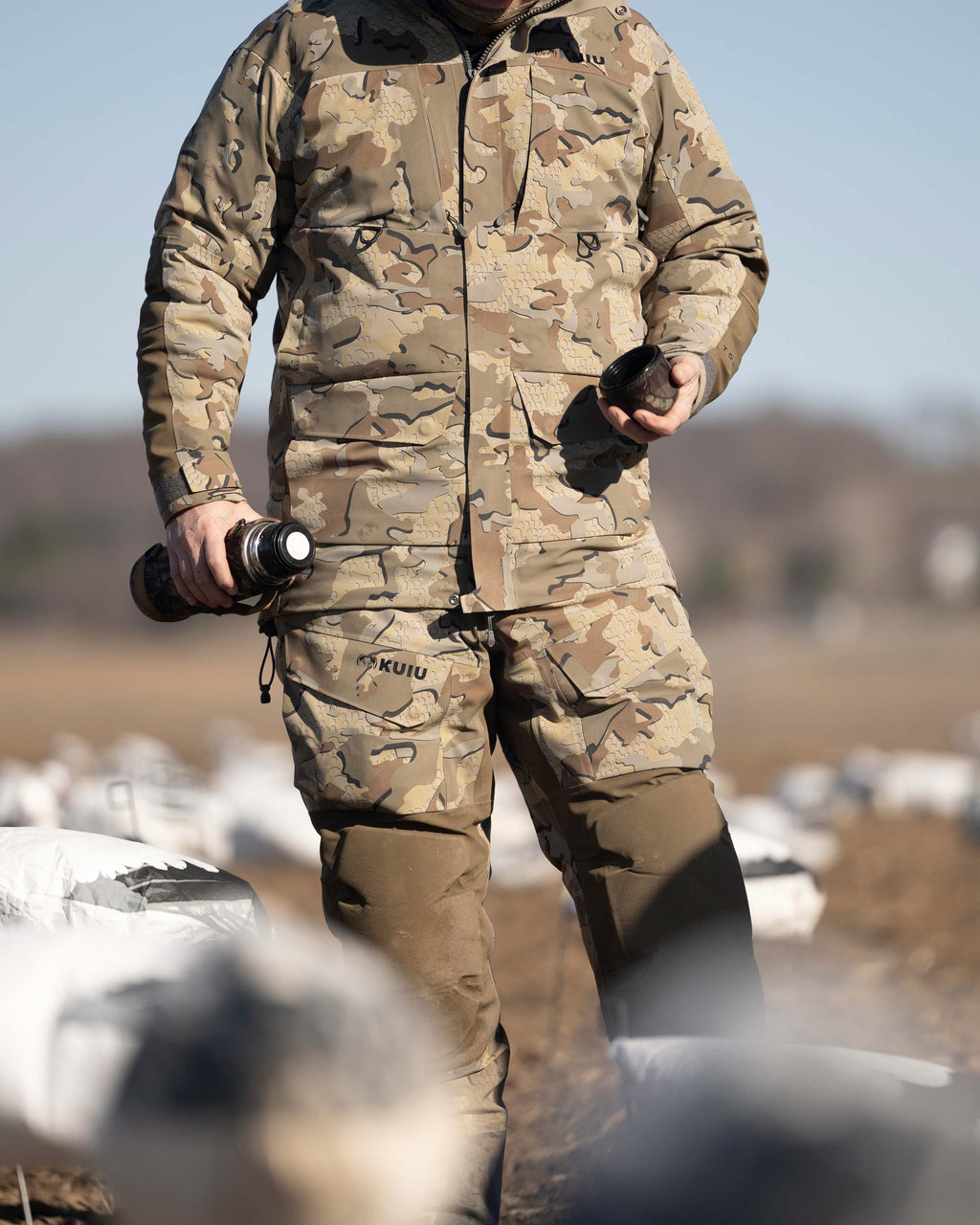 Waterfowl Hunter standing in a field of goose decoys, holding an open thermos, wearing Paragon Storm Shell Parka and Bib in Valo Camouflage