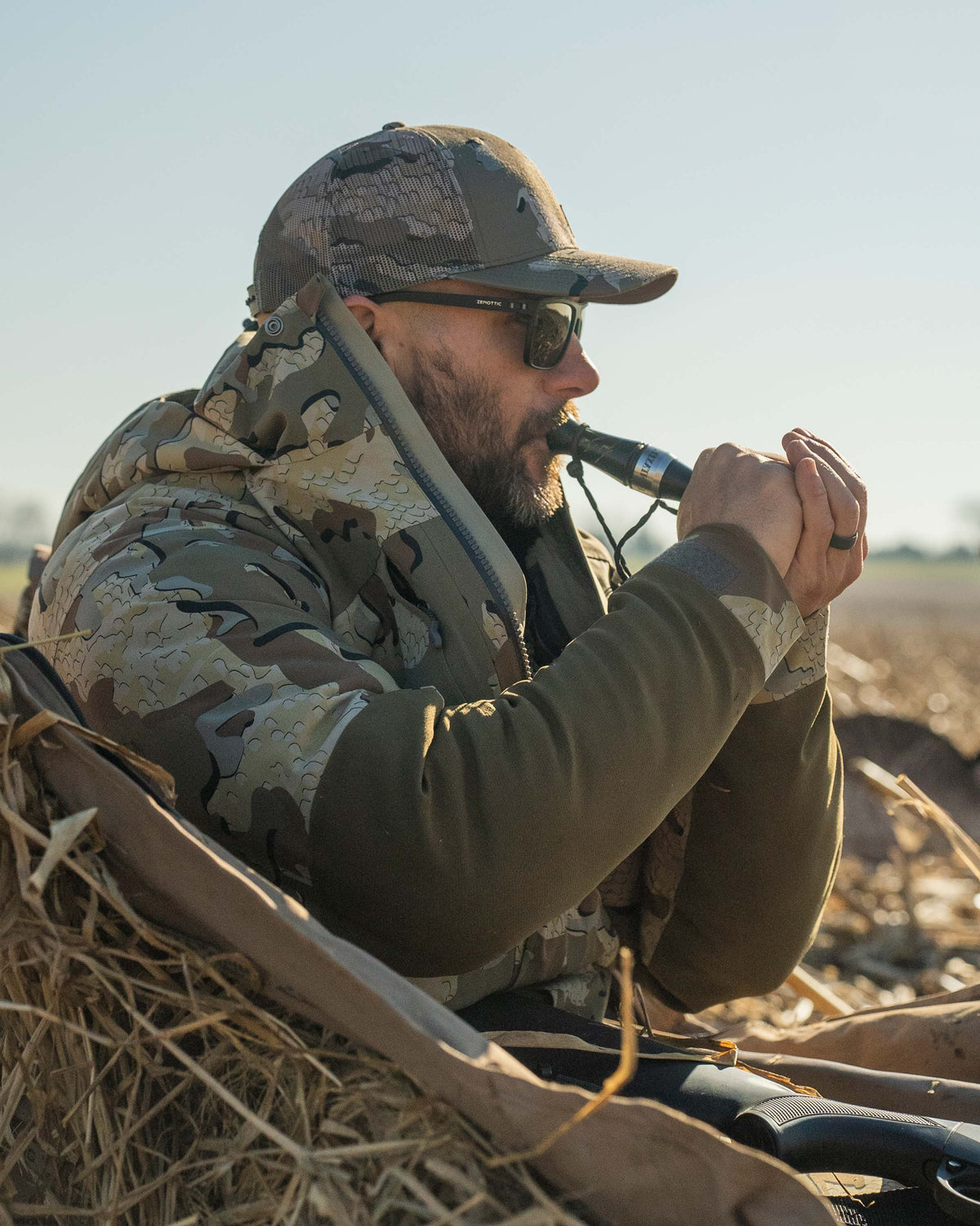 Side profile of Waterfowl Hunter sat propped in a field, calling ducks, wearing Paragon Storm Shell Parka in Valo Camouflage