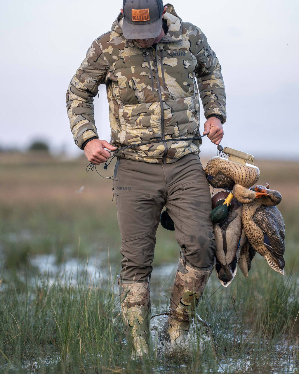 Hunter walking through a wet grass field wearing Reed Pant in Ash Brown carrying a half-dozen of duck decoys