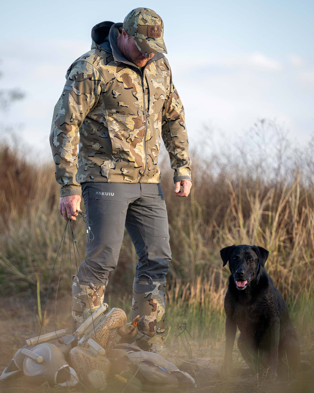 Hunter wearing Reed Pant in Gunmetal Grey in front of a grassy field with duck decoys standing next to a black labrador retriever dog