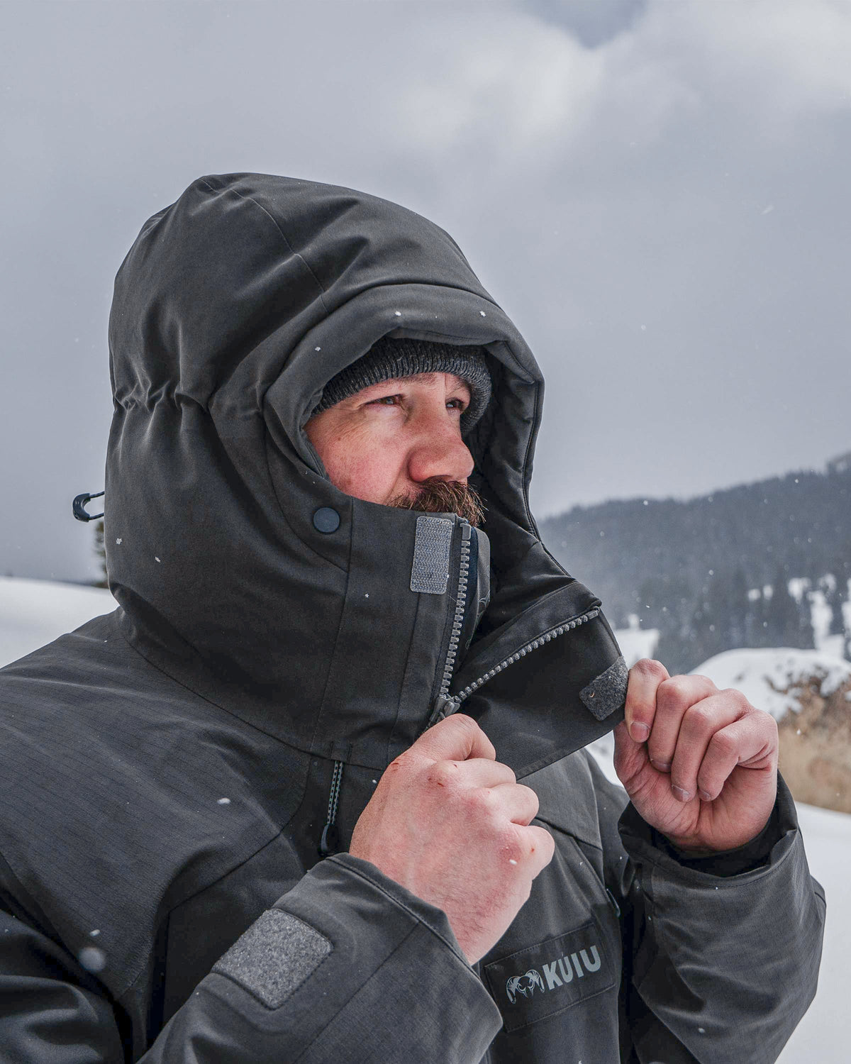 Hunter in a cold outdoor setting unzipping Super Down Arktuk Parka in Asphalt Grey, lowering the top chin guard