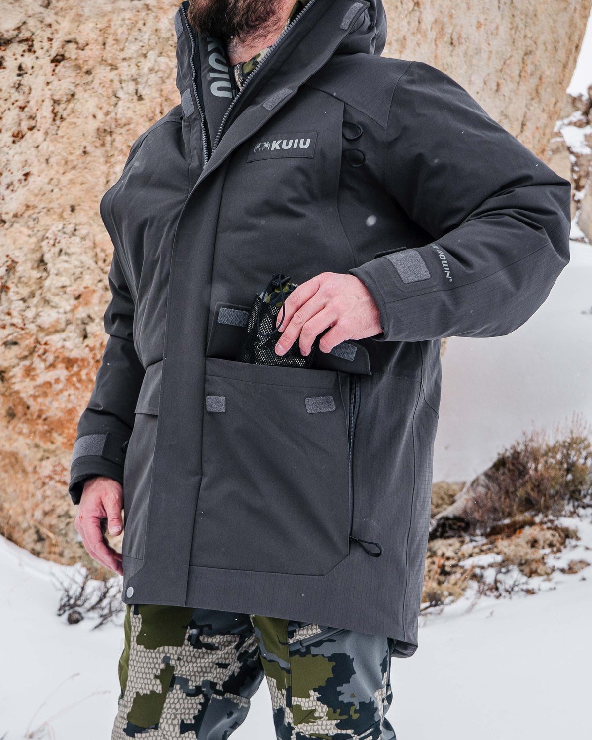 Hunter wearing Super Down Arktuk Parka in Asphalt Grey, standing in a snowy outdoor setting, putting a pair of gloves in his front cargo pockets featuring hook-and-loop closure with side gussets 