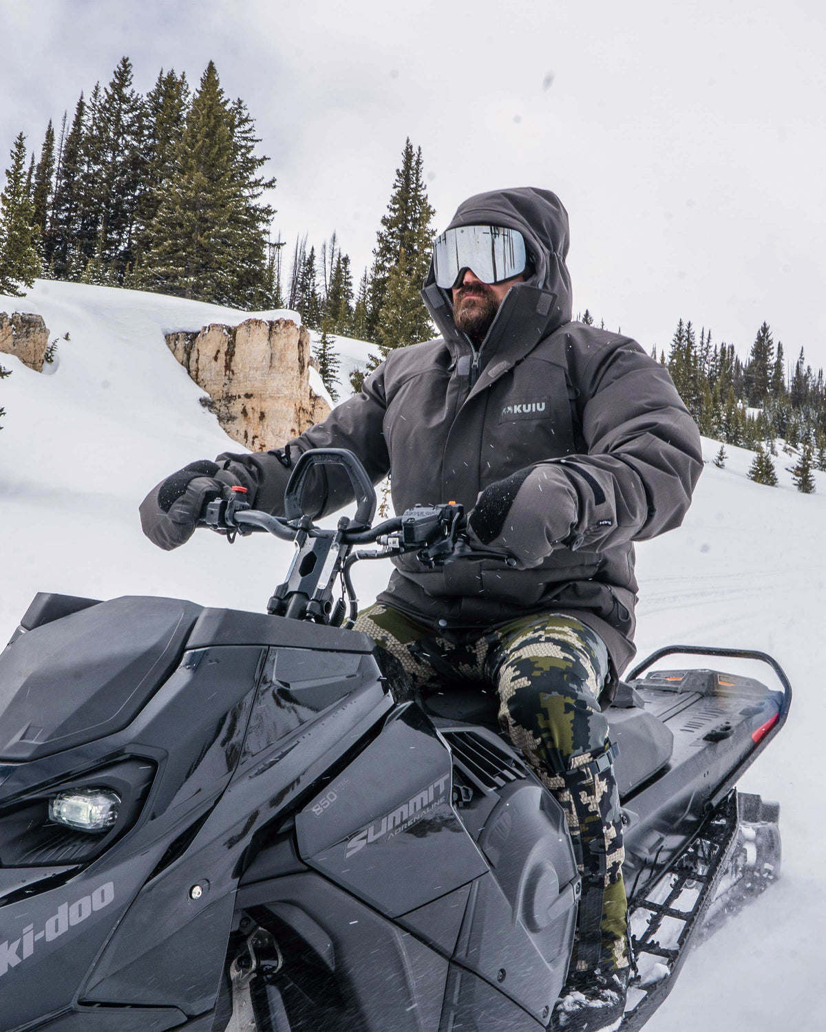 Hunter wearing Snow Down Arktuk Parka in Asphalt Grey, in a snowy environment, riding a black snow mobile 