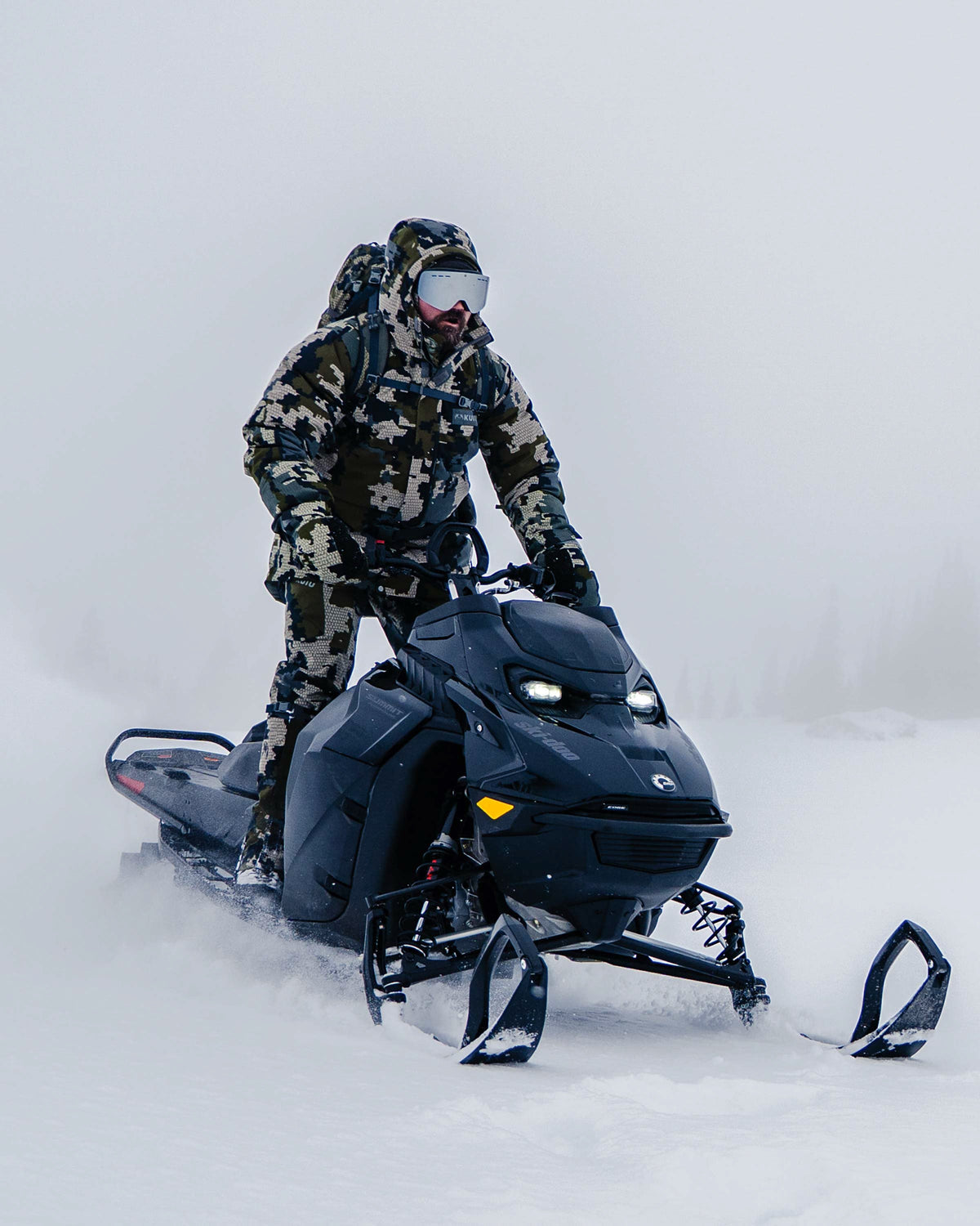 Hunter wearing Snow Down Arktuk Parka in Asphalt Grey riding a black snow mobile in a snowy landscape with low visibility