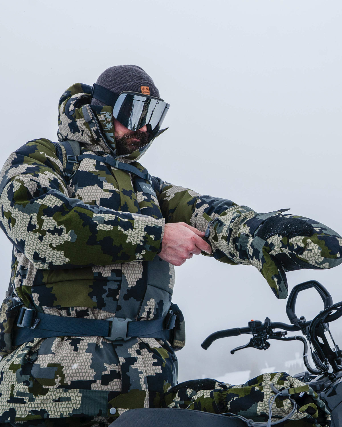 Hunter in a cold snowy condition wearing Super Down Arktuk Parka in Verde Camouflage putting on a pair of Super Down Arktuk Mitt in Verde Camouflag