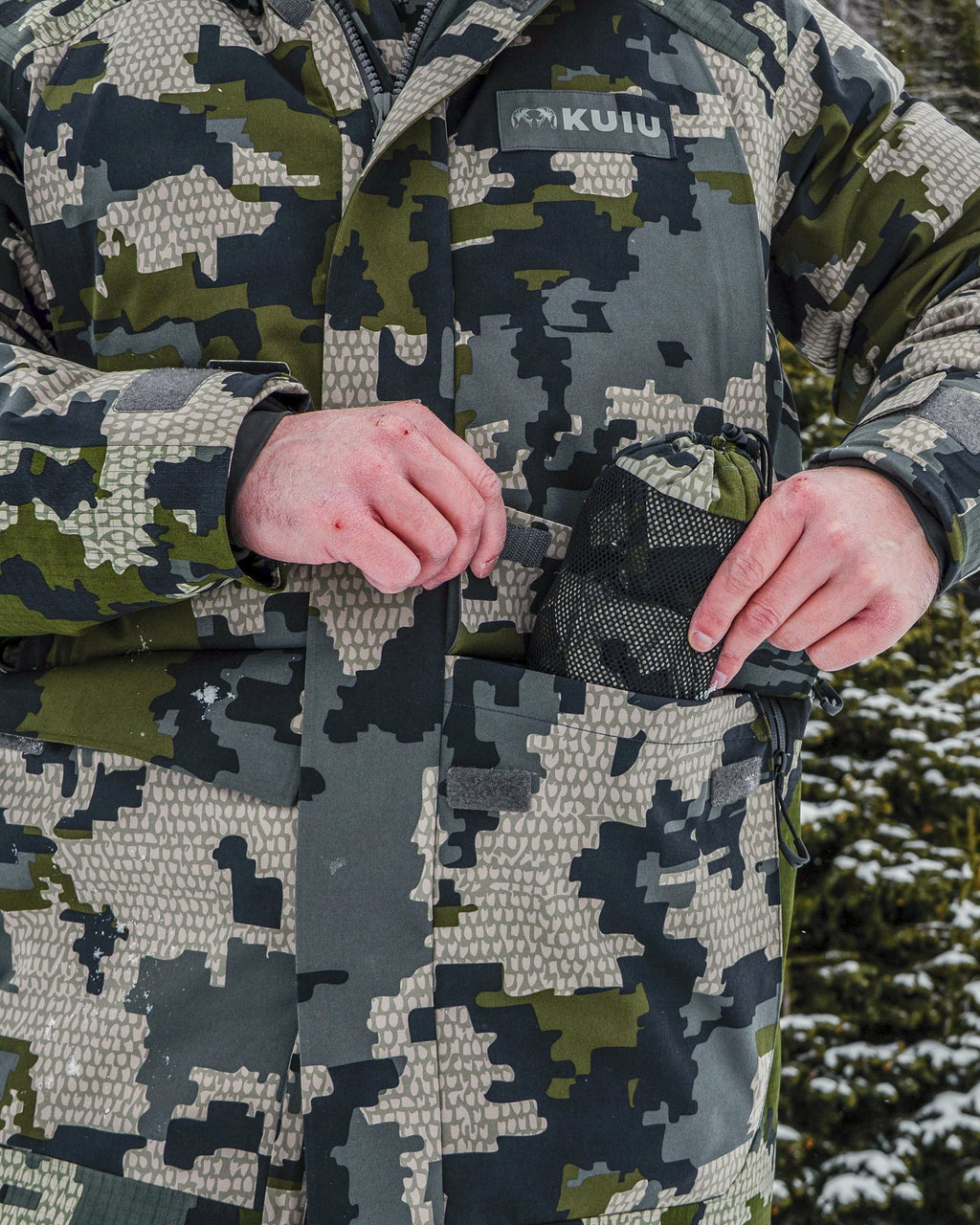 Close-up of hunter wearing Super Down Arktuk Parka in Verde Camouflage, putting a pair of gloves in his front cargo pockets featuring hook-and-loop closure