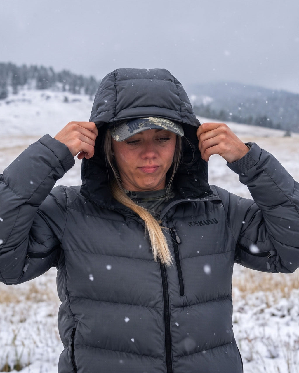 A woman in a snowy outdoor setting wearing a Super Down PRO Hooded Jacket in Gunmetal Grey adjusting the hood of her jacket as snow falls.