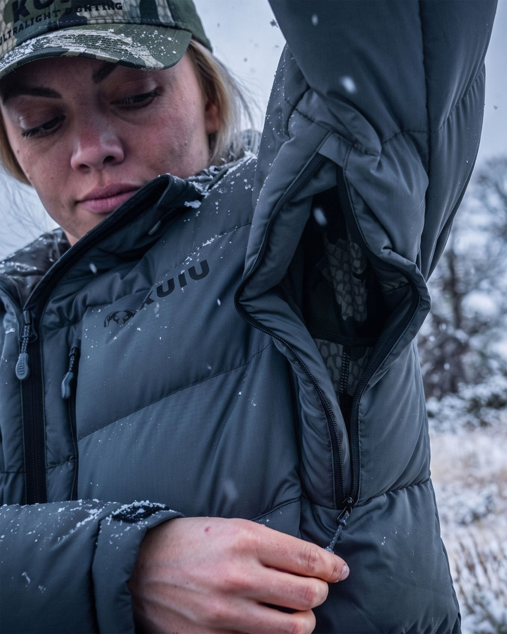 A woman wearing a Super Down PRO Hooded Jacket in Gunmetal Grey showing the underarm zippered pit vents, standing outdoors in a snowy landscape. 