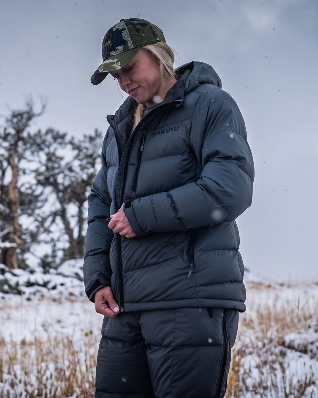 A woman wearing Super Down PRO Hooded Jacket and matching pants stands in a snowy outdoor landscape, looking down while adjusting the front zipper of her jacket.