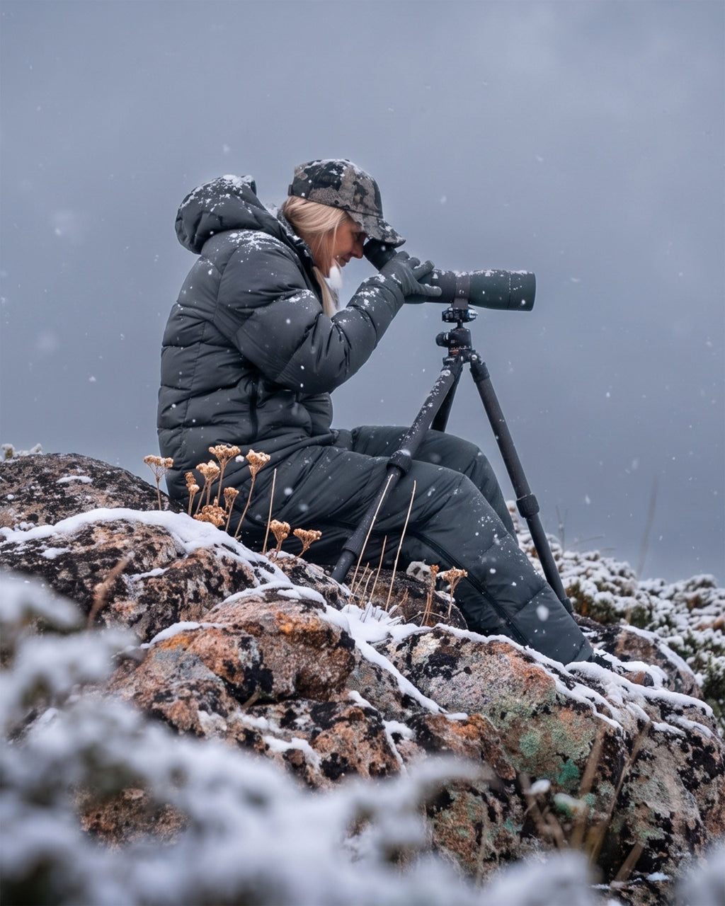 Woman wearing Super Down PRO Jacket and matching pant in Gunmetal Grey sits on a snowy rock, using a spotting scope mounted on a tripod in a snowy outdoor setting.