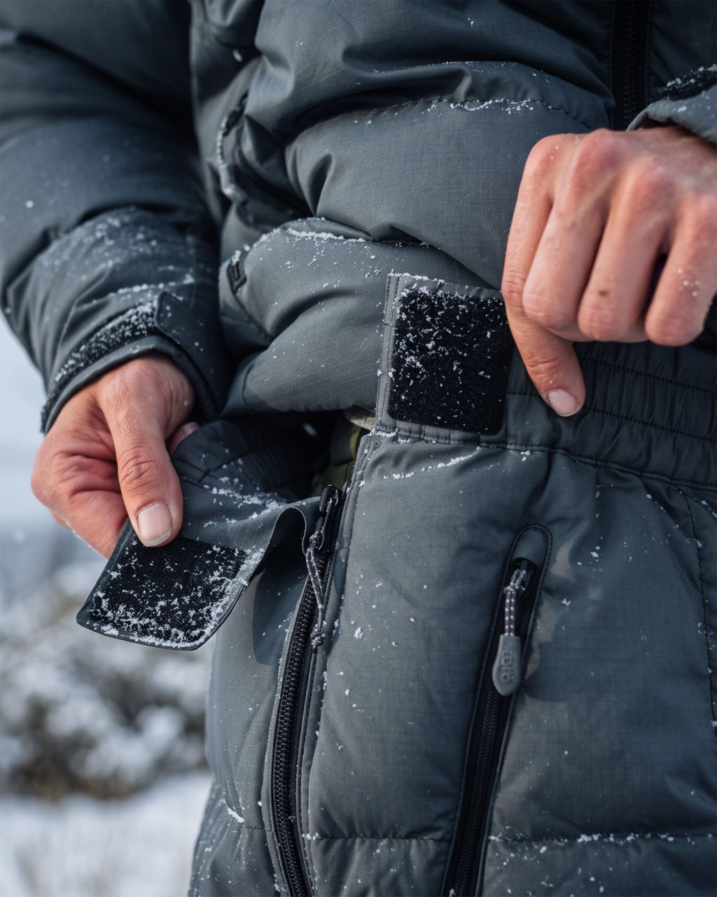 Close-up of a person wearing Women's Super Down Jacket and matching pant in Gunmetal Grey outdoors in the snow, opening a hook and loop tab closure at side waist.
