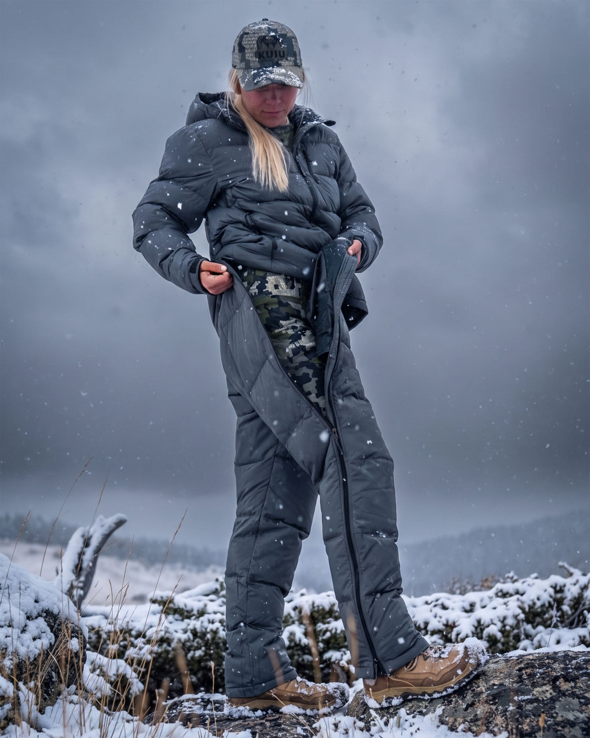 A woman wearing Super Down PRO Jacket and matching pant in Gunmetal Grey, standing outdoors in the snow under a cloudy gray sky.