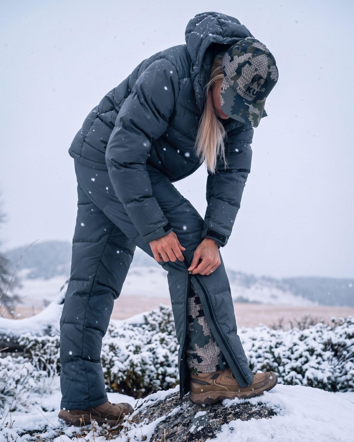 A woman in Super Down PRO Jacket and matching pant in Gunmetal Grey is bending down in a snowy outdoor setting.