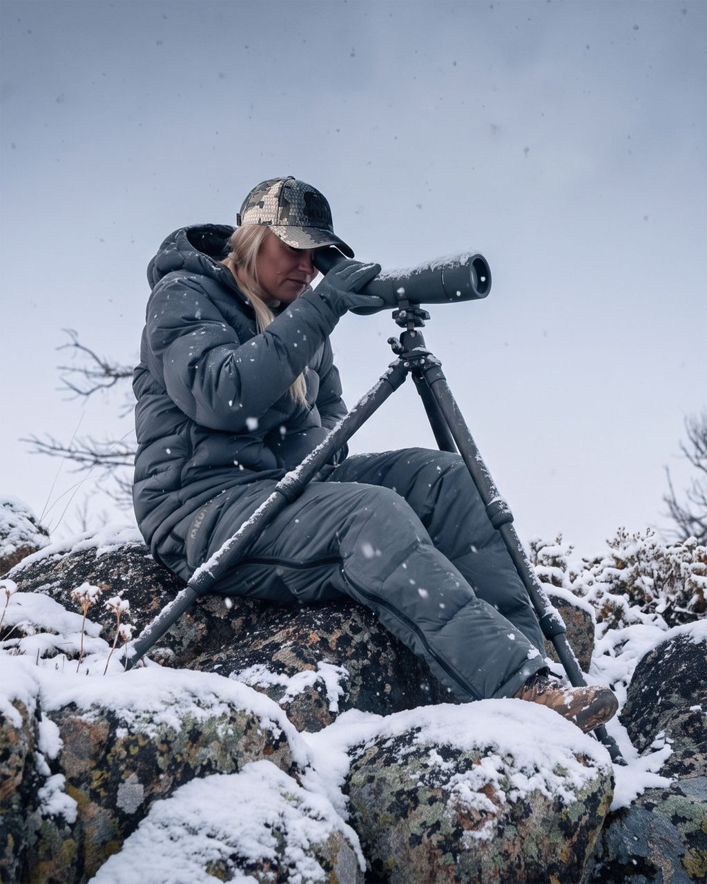 Woman in a winter outdoor setting wearing Super Down PRO Jacket and matching pant in Gunmetal Grey sits on a snow-dusted rock, using a spotting scope mounted on a tripod in a snowy outdoor setting.