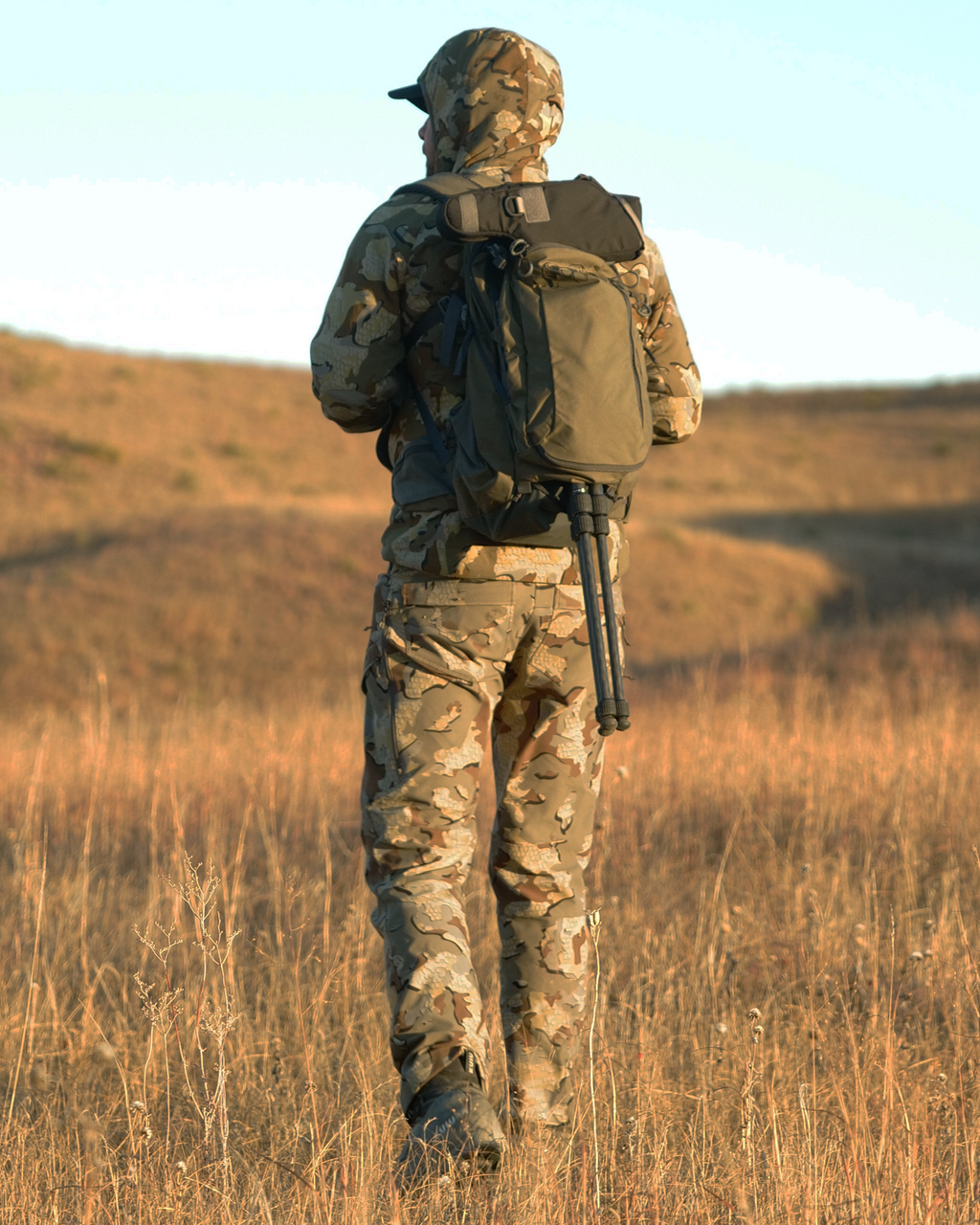 Hunter in Valo Camouflage wearing a Divide 1500 Packpack in Ash shown carrying a spotting scope attached to a tripod with the tripod legs fully extended and passed through the zippered divide pocket of the backpack