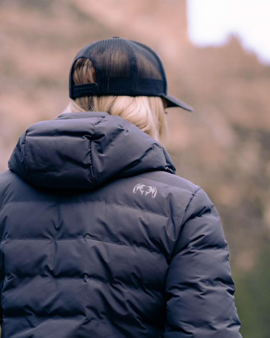 Back of woman wearing Elements Hooded Jacket in Gunmetal with KUIU Ram Logo on upper back right shoulder.